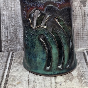 May include: A cylindrical ceramic candle holder with a dark blue and green mottled glaze, featuring vertical cutouts along the side. A metal ruler placed in front shows the object stands approximately 4.5 inches tall at its highest point.