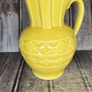 May include: A bright yellow ceramic pitcher featuring a flared spout, a curved handle, and a textured diamond pattern around the midsection. A metal ruler is positioned at the base, showing the pitcher stands approximately 8.5 inches tall.