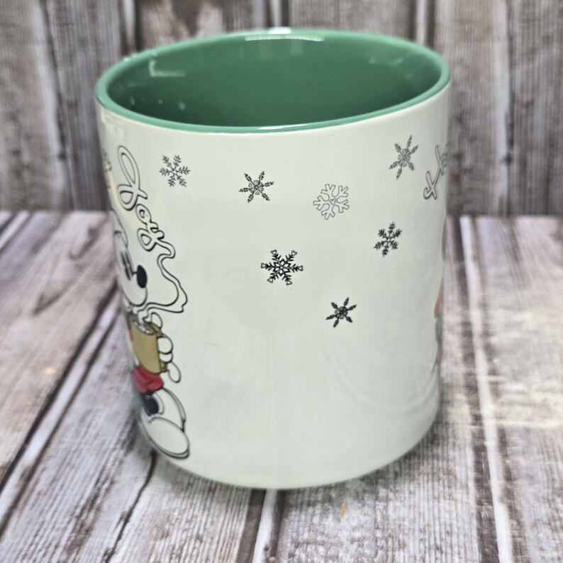 K&ouml;nnte beinhalten: Wei&szlig;e Keramiktasse mit gr&uuml;nem Innenraum und einem Cartoon-Bild von Mickey Mouse, der eine Tasse Kaffee h&auml;lt. Die Tasse ist mit Schneeflocken und dem Wort "Joy" in Schreibschrift verziert.