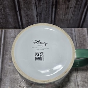 K&ouml;nnte beinhalten: Eine wei&szlig;e Disney-Tasse aus Keramik mit einem gr&uuml;nen Henkel. Der Boden der Tasse zeigt das Disney-Logo, den Text "Dishwasher and Microwave Safe Made in China" und das Zrike Brands-Logo.