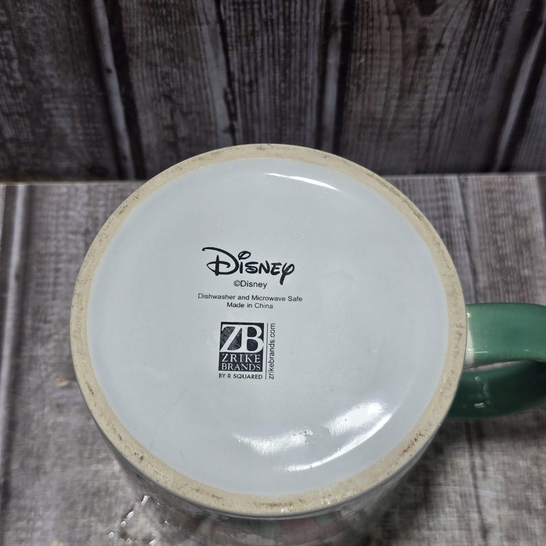 K&ouml;nnte beinhalten: Eine wei&szlig;e Disney-Tasse aus Keramik mit einem gr&uuml;nen Henkel. Der Boden der Tasse zeigt das Disney-Logo, den Text "Dishwasher and Microwave Safe Made in China" und das Zrike Brands-Logo.
