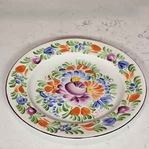 May include: A white ceramic plate with a hand-painted floral design. The plate features a central arrangement of purple, blue, orange, and yellow flowers, surrounded by a border of similar floral motifs. The rim is outlined in brown.