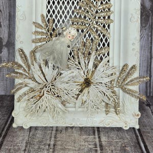 May include: A white ornate rectangular frame featuring a central diamond-patterned mesh grid. The frame is decorated with a small porcelain angel figurine wearing a sheer white dress, surrounded by white and gold glittered floral accents.