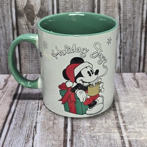 K&ouml;nnte beinhalten: Wei&szlig;er Keramikbecher mit gr&uuml;nem Henkel und Innenraum. Der Becher zeigt Mickey Mouse mit Weihnachtsm&uuml;tze, der eine Tasse Kakao h&auml;lt, mit den Worten "Holiday Joy" und Schneeflocken. Eine gr&uuml;ne Geschenkbox mit roter Schleife.