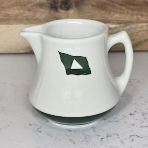 May include: A small, white ceramic creamer with a dark green base and a handle. A dark green flag-shaped design with a white triangle is on the front. The creamer has a spout for easy pouring.
