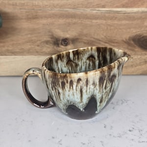 May include: A ceramic gravy boat with a handle and a spout. The exterior features a brown and cream glaze with a drip pattern. The interior is a light blue color. The gravy boat is on a white surface.