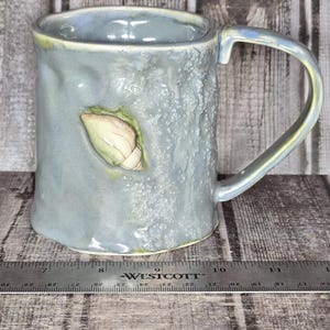 May include: A light blue ceramic mug with a textured, bumpy surface and a single handle. A small, cream-colored seashell is attached to the side. A metal Westcott ruler is positioned at the base, showing the mug is approximately 4.5 inches wide.