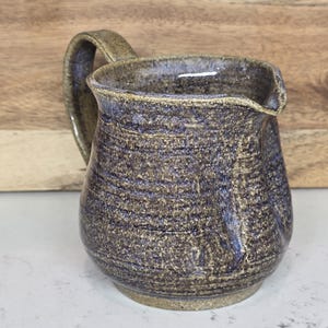 May include: A handmade ceramic pitcher with a handle. The pitcher has a textured surface with a speckled, earthy color palette of brown, blue, and tan. The pitcher is designed for serving beverages.