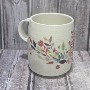 May include: Cream-colored ceramic mug with a floral design. The mug features a curved handle and a cylindrical shape. The floral pattern includes red, blue, and green leaves and flowers. The mug is suitable for hot or cold beverages.
