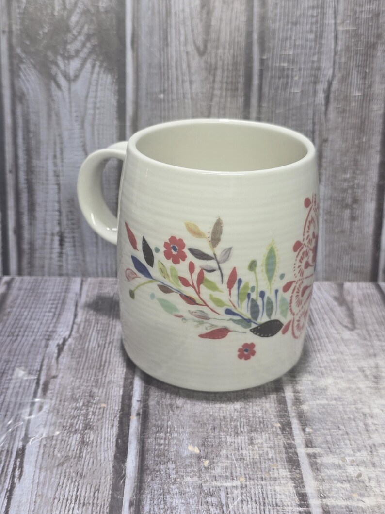 May include: Cream-colored ceramic mug with a floral design. The mug features a curved handle and a cylindrical shape. The floral pattern includes red, blue, and green leaves and flowers. The mug is suitable for hot or cold beverages.