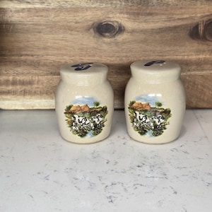 May include: A set of two ceramic salt and pepper shakers with a farmhouse theme. The shakers are beige with a brown roof and a green field with black and white cows.