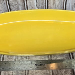 May include: A bright yellow rectangular ceramic baking dish with rounded corners and integrated handles. A metal Westcott ruler is positioned along the bottom edge, showing the dish measures approximately 14 inches in total length.