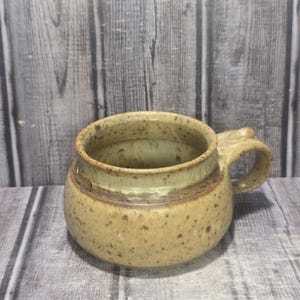 May include: A speckled, handmade ceramic mug with a rounded body and a small handle. The mug features a light brown base with a lighter, greenish-tan rim and handle. The mug is designed for hot beverages.
