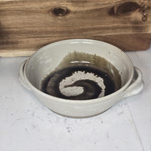 May include: A white ceramic bowl with two handles. The bowl has a brown glaze with a swirl pattern.
