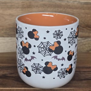 May include: White ceramic mug with an orange interior. The exterior is decorated with black Mickey Mouse silhouettes with orange bows, spiderwebs, bats, and stars. The mug is a Halloween-themed item.