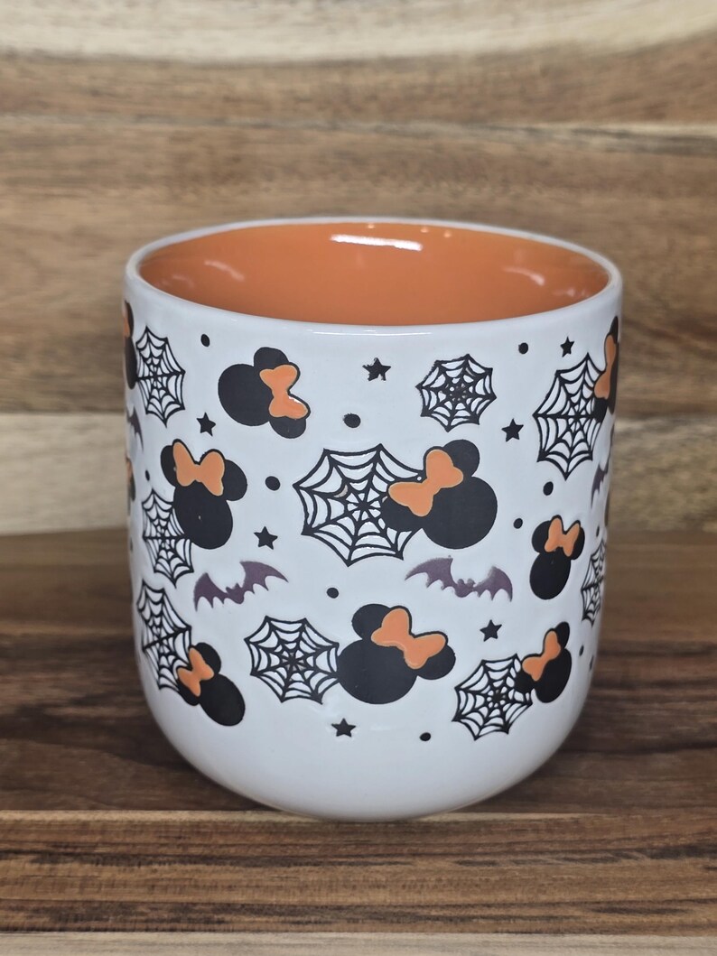 May include: White ceramic mug with an orange interior. The exterior is decorated with black Mickey Mouse silhouettes with orange bows, spiderwebs, bats, and stars. The mug is a Halloween-themed item.