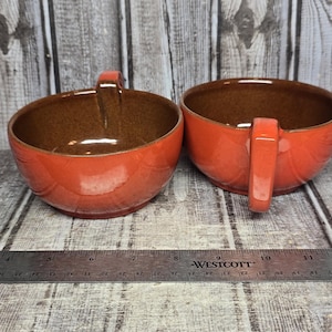 May include: Two ceramic soup bowls with a glossy orange exterior and a speckled brown interior, each featuring a single handle. A metal Westcott ruler is positioned in front, showing the bowls have a diameter of approximately 5 inches.