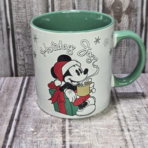 K&ouml;nnte beinhalten: Wei&szlig;e Keramik-Tasse mit gr&uuml;nem Innenraum und Henkel. Die Tasse zeigt ein Cartoon-Bild von Mickey Mouse mit Weihnachtsm&uuml;tze, die eine Tasse Kakao h&auml;lt. Die Worte "Holiday Joy" stehen &uuml;ber der Figur, mit Schneeflocken.