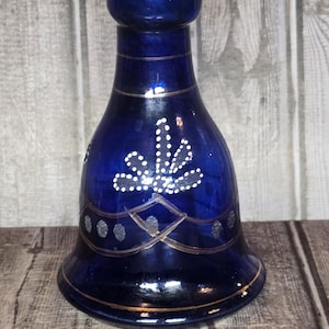 May include: A cobalt blue glass candle holder with a flared base and a cylindrical top. The glass features hand-painted silver horizontal lines, a central white dotted floral motif, and a row of silver circles. A Westcott ruler shows a 5-inch width.