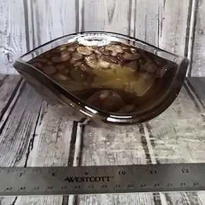 May include: A dark brown, translucent glass bowl with a curved, leaf-like shape. A metal Westcott ruler is positioned in front of the bowl, showing the object spans approximately 5 inches in width from its widest points.