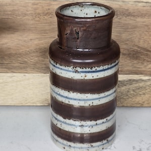 May include: A cylindrical ceramic vase with a dark brown top and horizontal stripes in brown, white, and light blue. The vase has a slightly textured surface and a small opening at the top. The vase is a decorative item.