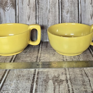 May include: Two matching mustard yellow ceramic soup mugs with wide, rounded bowls and square-shaped handles. A metal ruler is positioned in front of the mugs, showing that each mug measures approximately 5 inches in width from edge to handle.