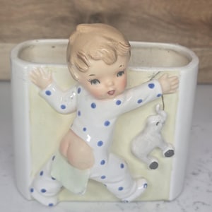 May include: A ceramic planter featuring a baby in a polka-dot onesie with arms outstretched. The baby is holding a small toy elephant. The planter has a rectangular shape with a light yellow interior and white exterior.