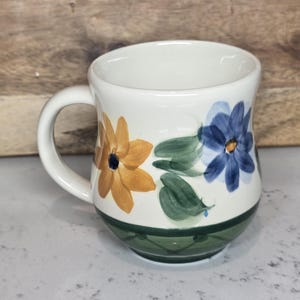 May include: White ceramic mug with a curved handle, decorated with hand-painted flowers in shades of yellow and blue, along with green leaves. The base of the mug features a green band with a diamond pattern.