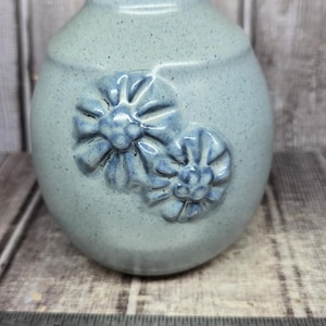 May include: A light blue speckled ceramic vase with two embossed flower designs on the front. A metal Westcott ruler is positioned at the base, showing the vase is approximately 3.5 inches wide at the point where it aligns with the ruler markings.