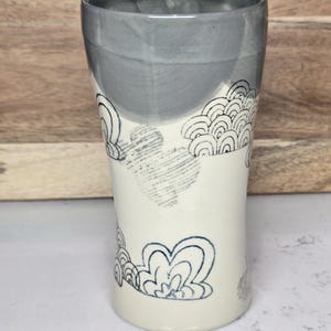 May include: A handmade ceramic tumbler with a gray top and a cream-colored body. The tumbler features a hand-drawn cloud design in dark blue. The design is repeated around the body of the tumbler. The tumbler is approximately 5 inches tall.