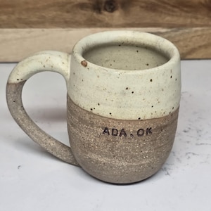 May include: A handmade ceramic mug with a speckled cream-colored top and a textured brown base. The handle is curved and matches the base. The text "ADA, OK" is stamped on the side. The mug is designed for hot beverages.