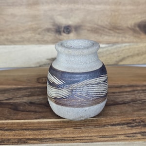 May include: A small, handcrafted ceramic vase with a rounded body and a narrow neck. The vase features a light brown base with a dark brown band and a decorative pattern of horizontal lines. The vase is displayed on a wooden surface.