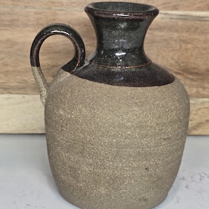 May include: A small, handcrafted ceramic pitcher with a handle. The pitcher has a textured, light brown body and a dark brown and black glazed top and handle. The design is simple and rustic, suitable for decorative use.