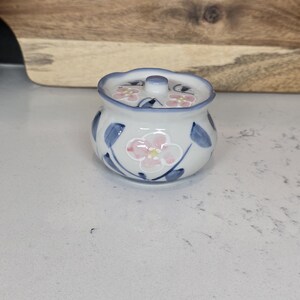 May include: A small, white ceramic jar with a lid, decorated with pink floral designs and blue leaves. The jar has a blue rim and a small knob on the lid. The design is hand-painted, giving it a unique, artisanal feel.
