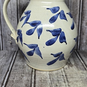 May include: A cream-colored ceramic pitcher with a speckled finish and a hand-painted pattern of dark blue leaf clusters. The pitcher features a curved handle on the left side, a wide rounded body, and a flared rim with a small pouring spout.