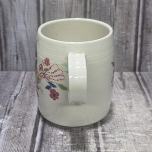 May include: Cream-colored ceramic mug with a white handle and floral design. The mug features a ribbed texture and is adorned with red, blue, and white floral accents. A classic and charming piece for enjoying hot beverages.