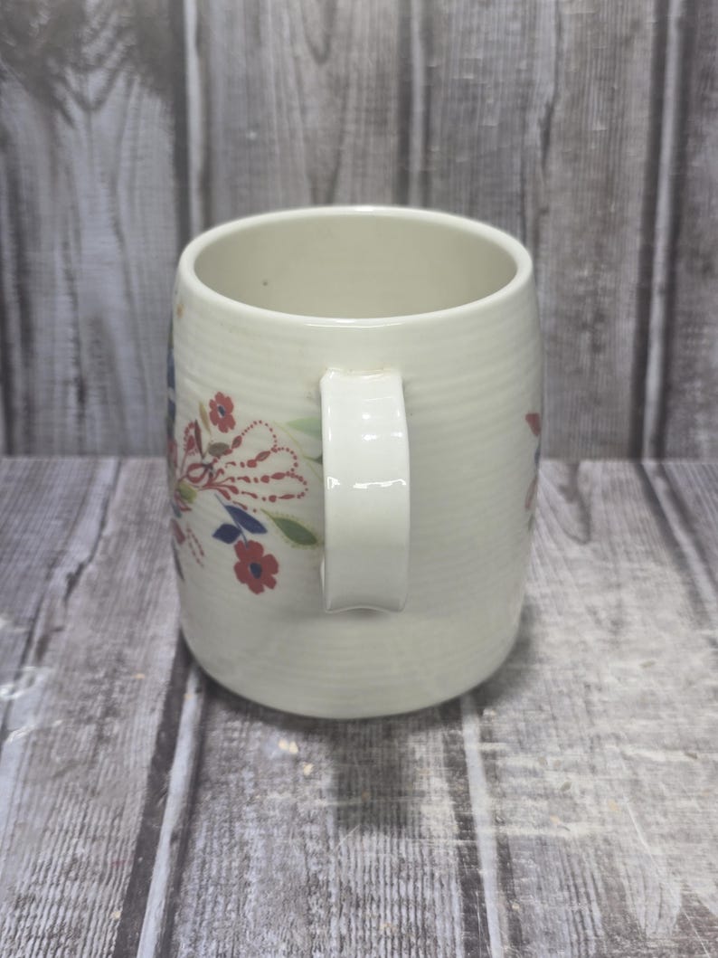 May include: Cream-colored ceramic mug with a white handle and floral design. The mug features a ribbed texture and is adorned with red, blue, and white floral accents. A classic and charming piece for enjoying hot beverages.