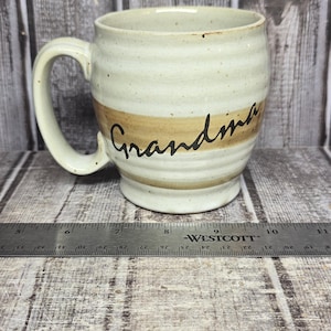 May include: A ceramic mug with a speckled off-white glaze and a horizontal tan band featuring the word Grandma in black script. The mug has a curved handle and a ribbed texture. A metal ruler shows the mug is approximately 4.5 inches wide.