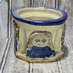 May include: A handmade ceramic planter with a blue rim and blue accents. The planter features a beige exterior with a blue figure design. The planter is a unique piece of art.