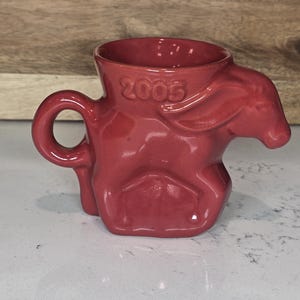May include: A red ceramic mug with a unique design, featuring a handle and a sculpted form resembling an animal. The year "2005" is embossed on the side. The mug is set against a neutral background.