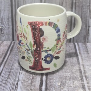 May include: Cream-colored ceramic mug with a large, colorful letter "P" and floral designs. The letter is primarily red with a textured pattern. The mug features a handle and is decorated with various flowers and leaves.