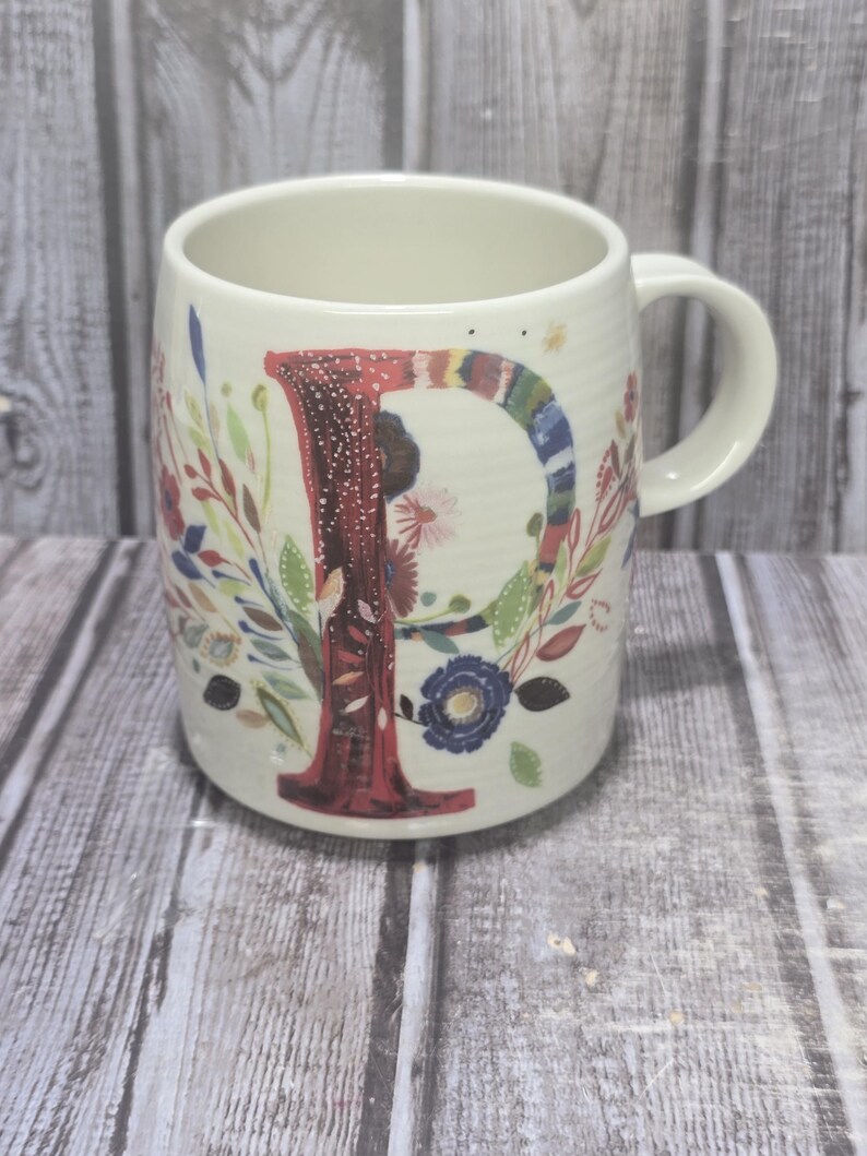 May include: Cream-colored ceramic mug with a large, colorful letter "P" and floral designs. The letter is primarily red with a textured pattern. The mug features a handle and is decorated with various flowers and leaves.