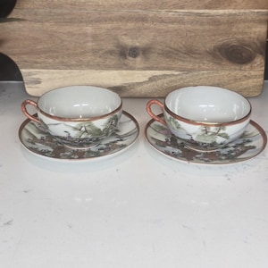 May include: Two white teacups and saucers with a landscape design. The cups have a gold rim and handle, and the saucers feature a detailed scene with trees and buildings. The set is placed on a white surface.