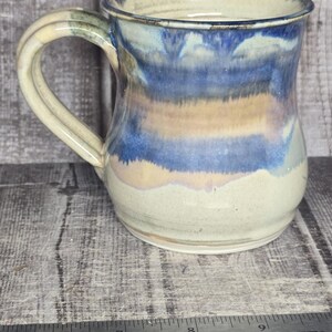 May include: A ceramic mug with a rounded body and a curved handle, featuring a glaze of cream, tan, and deep blue horizontal bands. A metal Westcott ruler is positioned in front, showing the mug stands approximately 4.5 inches in height.