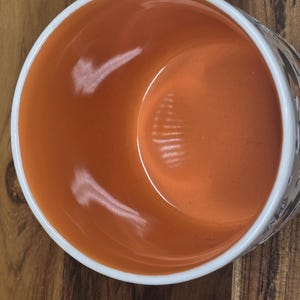 May include: A ceramic bowl with an orange interior and a white rim. The exterior features a decorative pattern. The bowl is set on a wooden surface. The bowl is empty.