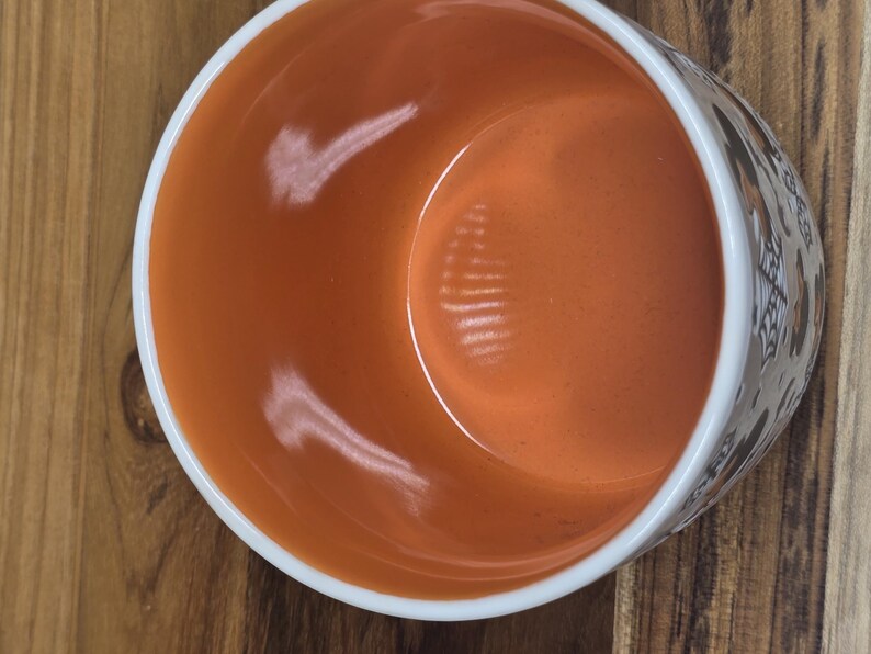 May include: A ceramic bowl with an orange interior and a white rim. The exterior features a decorative pattern. The bowl is set on a wooden surface. The bowl is empty.