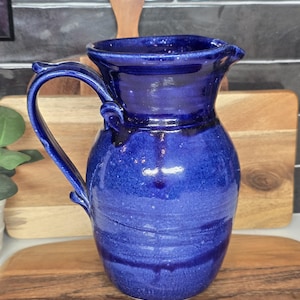 May include: A handmade, cobalt blue ceramic pitcher with a curved handle and a pouring spout. The pitcher has a glossy finish and visible texture from the pottery process. The pitcher is displayed on a wooden cutting board.
