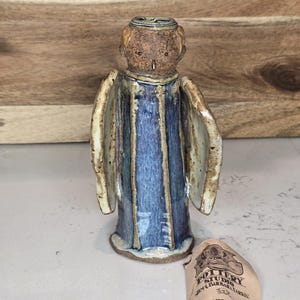 May include: A ceramic angel figurine with a blue and brown color scheme. The angel has a detailed face and wings, and is wearing a blue robe with vertical stripes. A tag with text is visible.