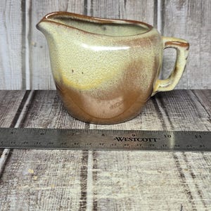 May include: A ceramic creamer pitcher with a two-tone glaze featuring a light cream upper section and a dark brown lower section. The pitcher has a curved handle and a pouring spout. A metal ruler placed in front shows a width of about 6 inches.