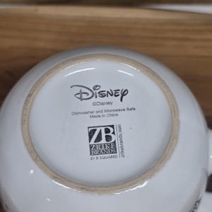 May include: White ceramic Disney mug with a black and white graphic. The bottom of the mug features the Disney logo, along with text indicating it is dishwasher and microwave safe, and the Zrike Brands logo. Made in China.